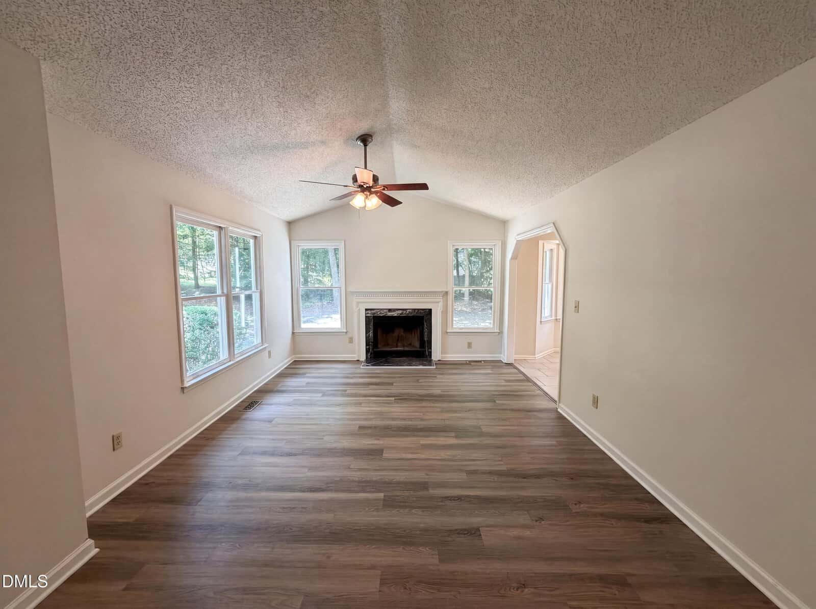 A bright living room with light-colored walls, wood flooring, large windows, a ceiling fan with lights, and a fireplace centered on the far wall. Natural light streams in from the left.
