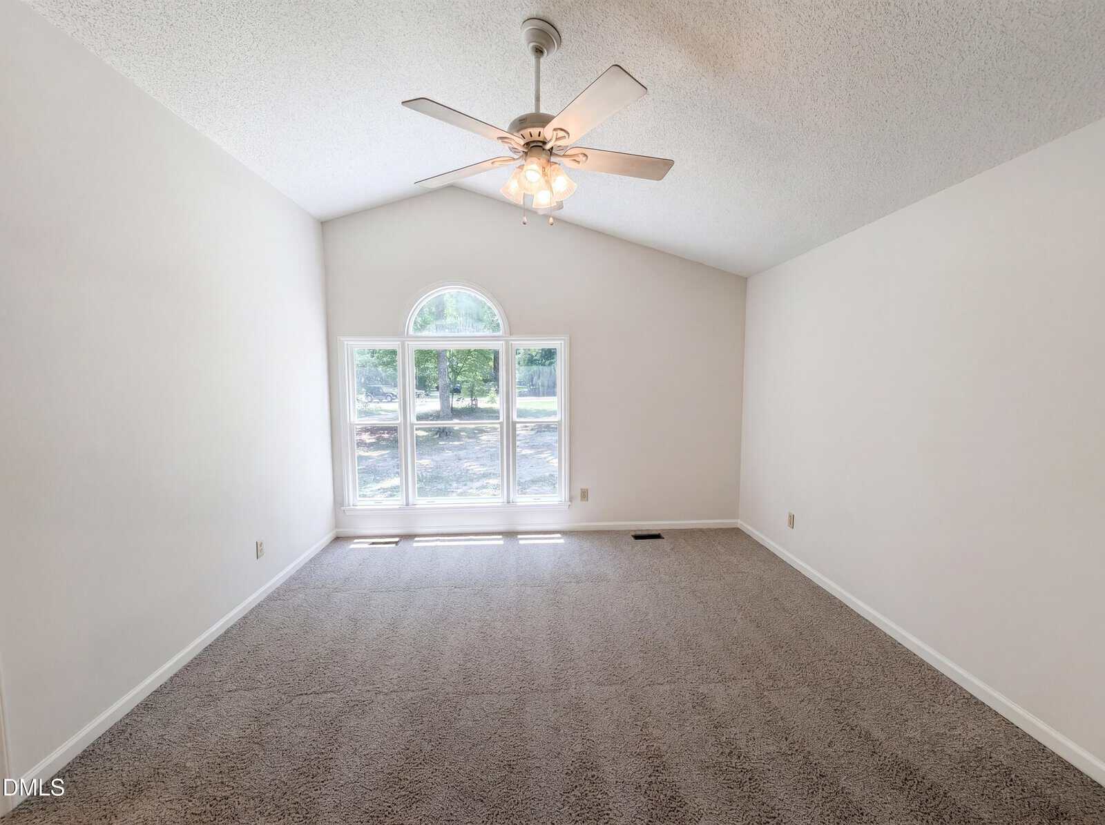 Empty room with beige carpet, white walls, a vaulted ceiling, ceiling fan with lights, and a large window with a curved top letting in natural light and offering a view of trees outside.