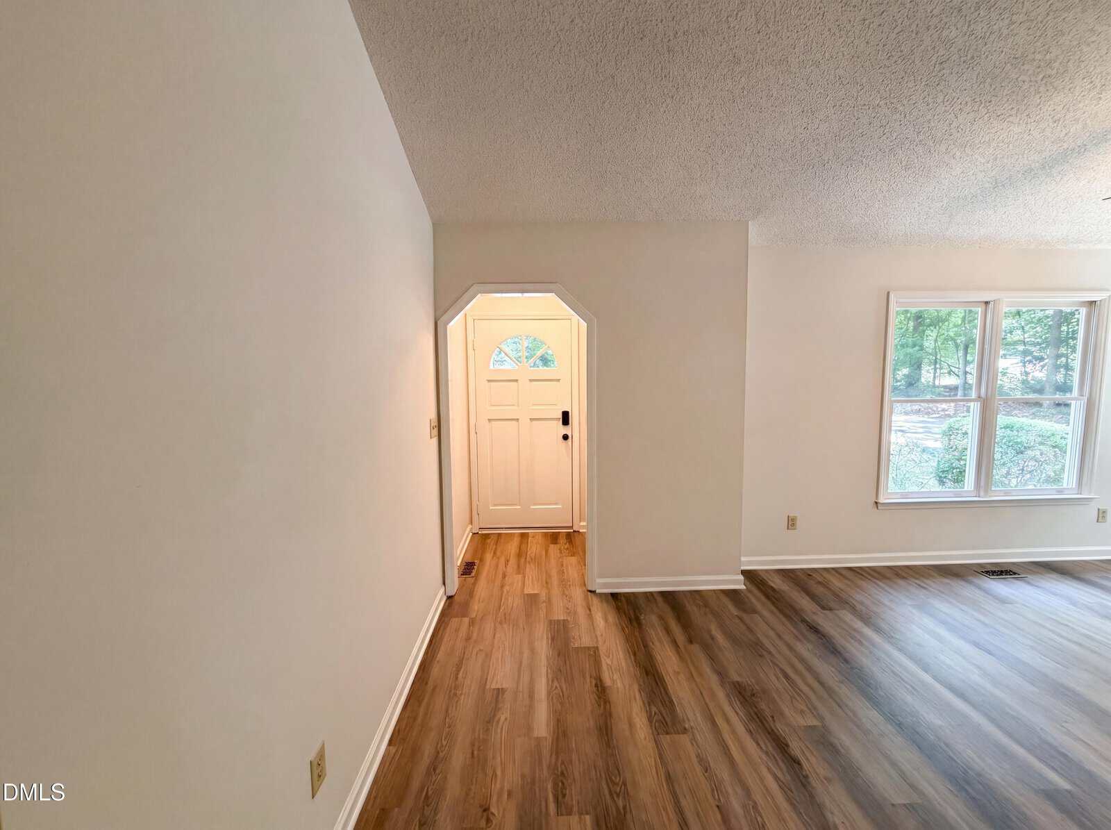 A bright entryway with a white front door opens into a spacious room featuring large windows and wood-style flooring; the walls are painted a light color, and natural light fills the space.