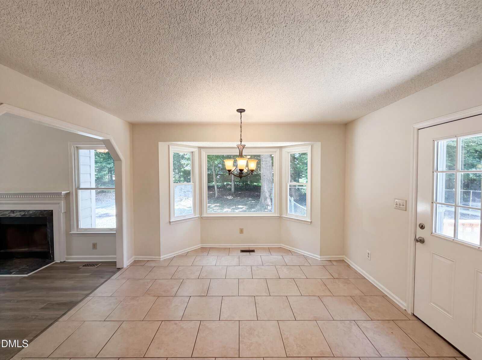A bright, empty dining area with tiled floors, a bay window overlooking a green yard, a hanging chandelier, and a white door. An archway leads to a room with a fireplace on the left.
