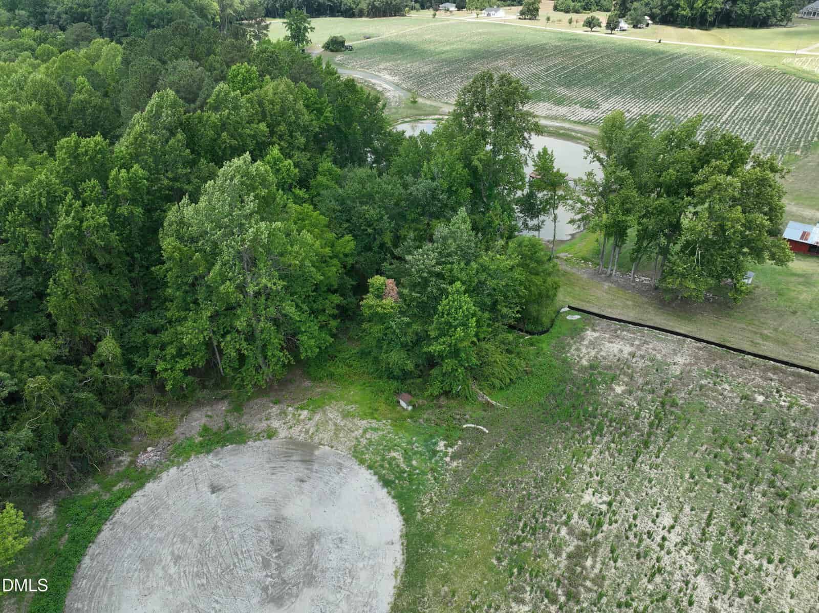 Aerial view of a rural landscape with dense green trees, a small pond, open fields, a circular dirt area in the foreground, and a red-roofed structure near the tree line.