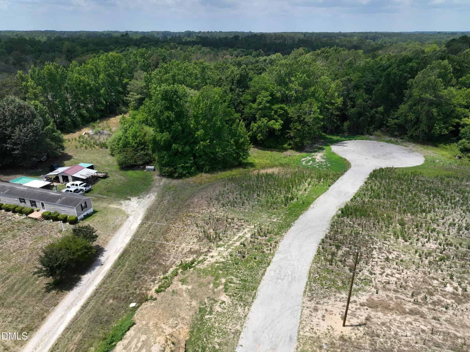 Aerial view of a rural area with a white house, several outbuildings, a gravel driveway, small plants or crops, and dense forest in the background under a partly cloudy sky.