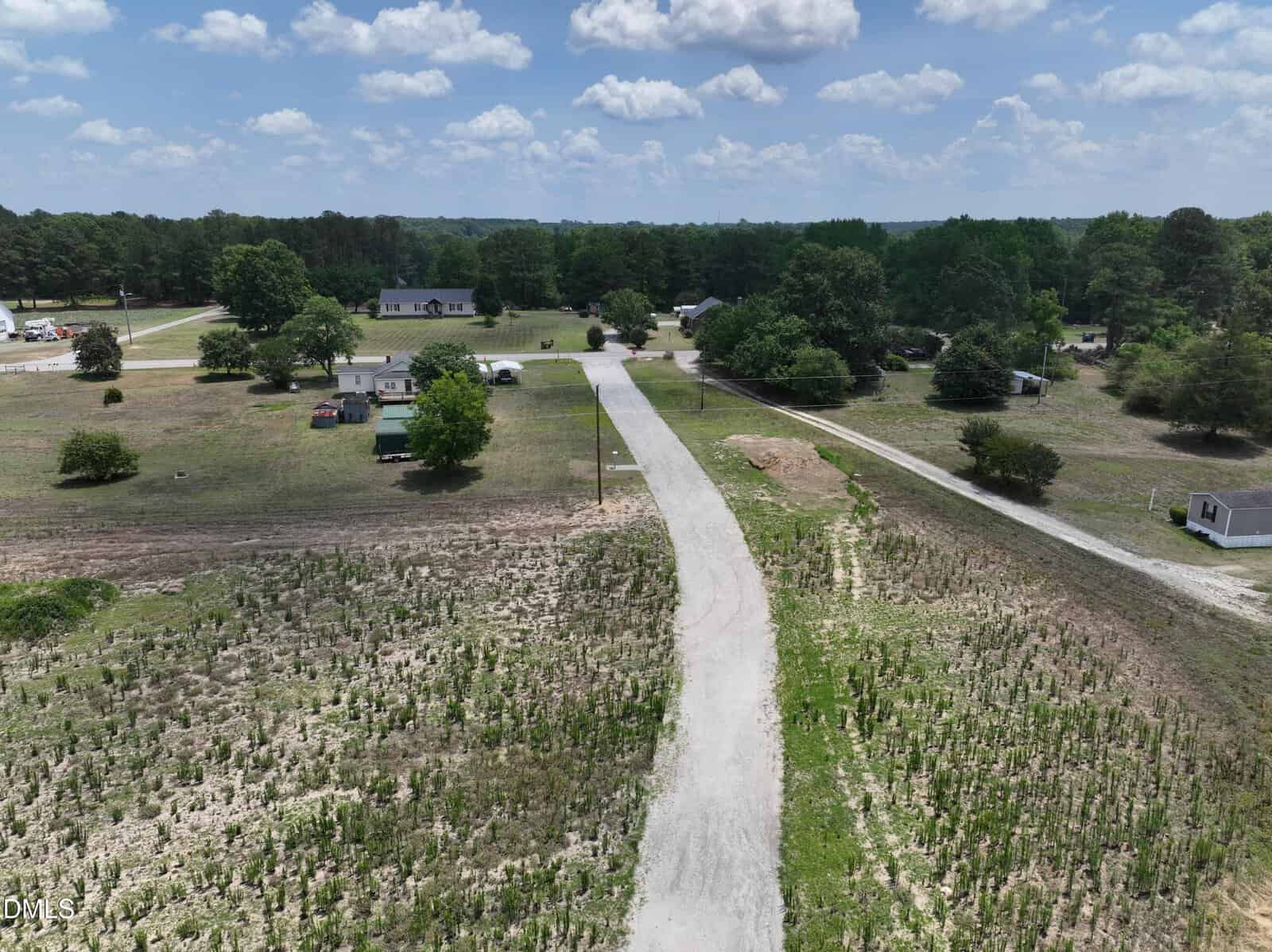 A narrow paved road winds through a rural area with scattered houses, trees, patches of grass, and clear blue skies with scattered clouds overhead.