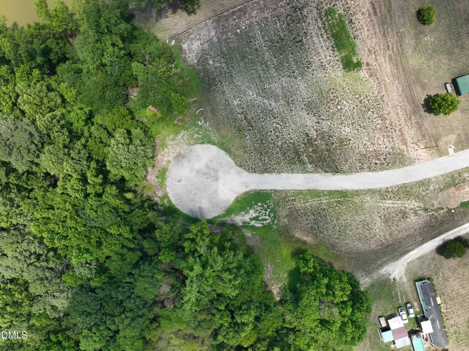 Aerial view of a rural cul-de-sac surrounded by trees and grass, with dirt roads and a few buildings visible on the right side of the image.
