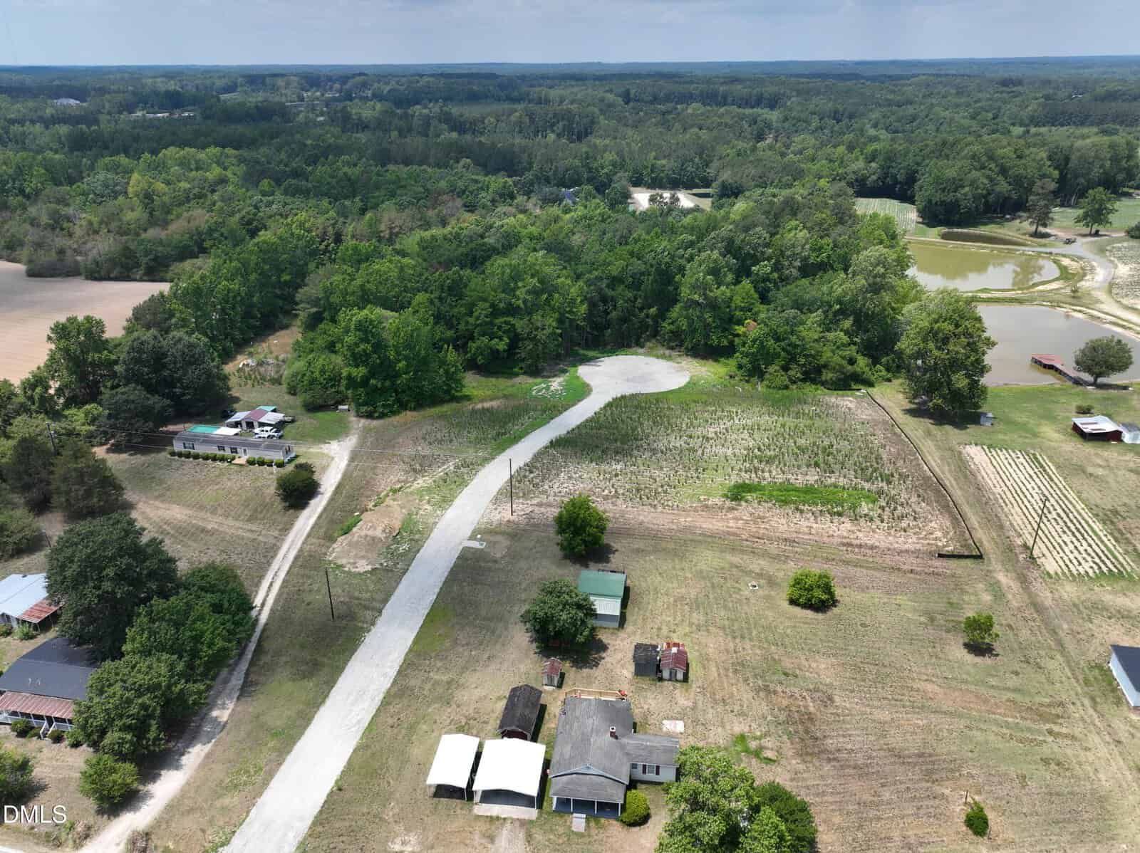 Aerial view of a rural area with a curved white road, scattered houses, green trees, and fields. Some small buildings and a pond are visible, surrounded by open land and forested areas.