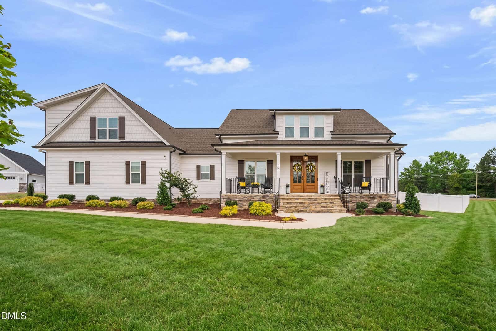 A modern, single-story house with white siding, stone accents, and a covered porch at 15 Old Garden Lane in Youngsville, featuring manicured landscaping and lush greenery under a bright blue sky.