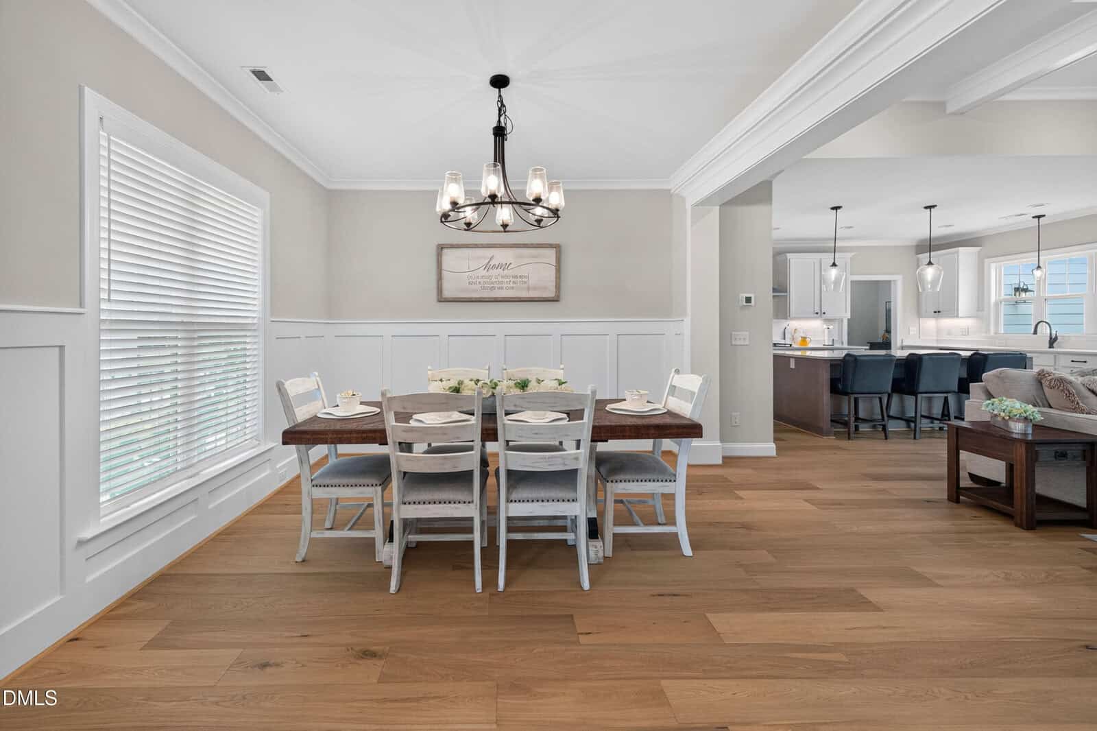 Bright dining room with a wooden table set for six, white chairs, a modern chandelier, and large window. Open layout leads to a kitchen with barstools and a glimpse of a living area.