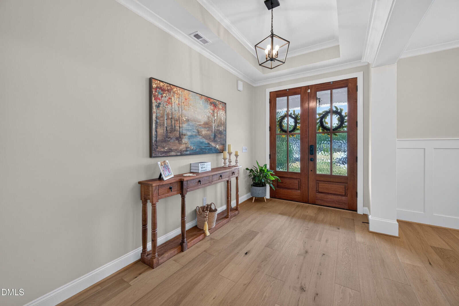 Bright entryway with double wooden doors, a console table holding candles, a basket, a framed photo, a plant, and wall art depicting a wooded path. Light wood floors and beige walls create a welcoming atmosphere.