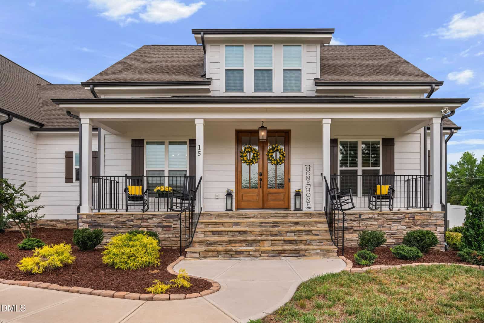 A modern white house with a covered front porch, stone steps, double wooden doors with wreaths, black railings, yellow chairs, and neatly landscaped bushes and shrubs.