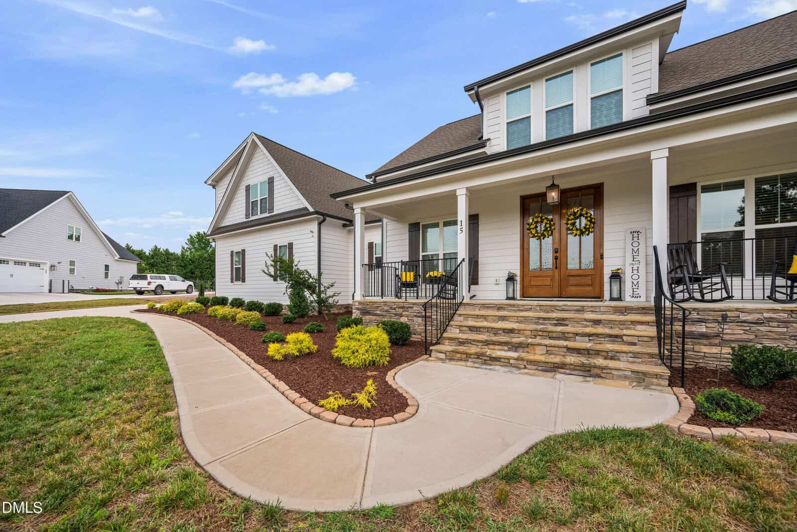 A modern suburban house with white siding, a covered front porch, double wooden doors, stone steps, and neatly landscaped yard with shrubs and flowers lining a curved sidewalk.