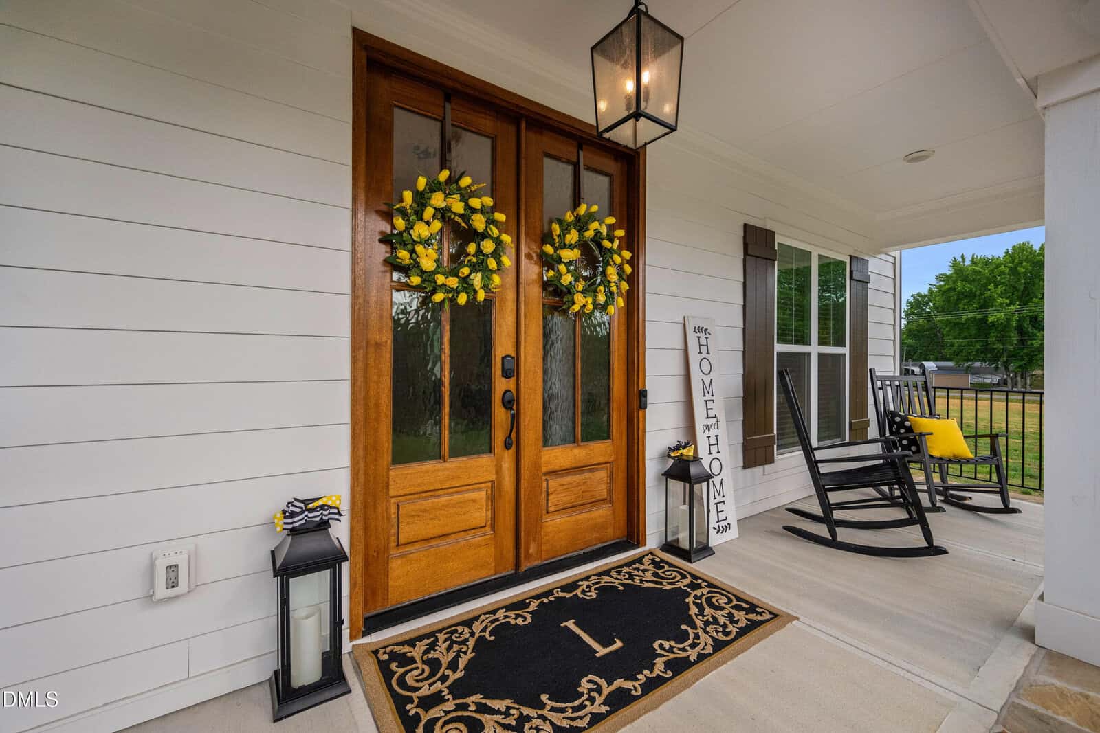 A front porch with double wooden doors, each decorated with a yellow flower wreath. Two black rocking chairs and a "HOME" sign sit nearby; a monogrammed doormat and lanterns are on the floor. The porch is clean and inviting.