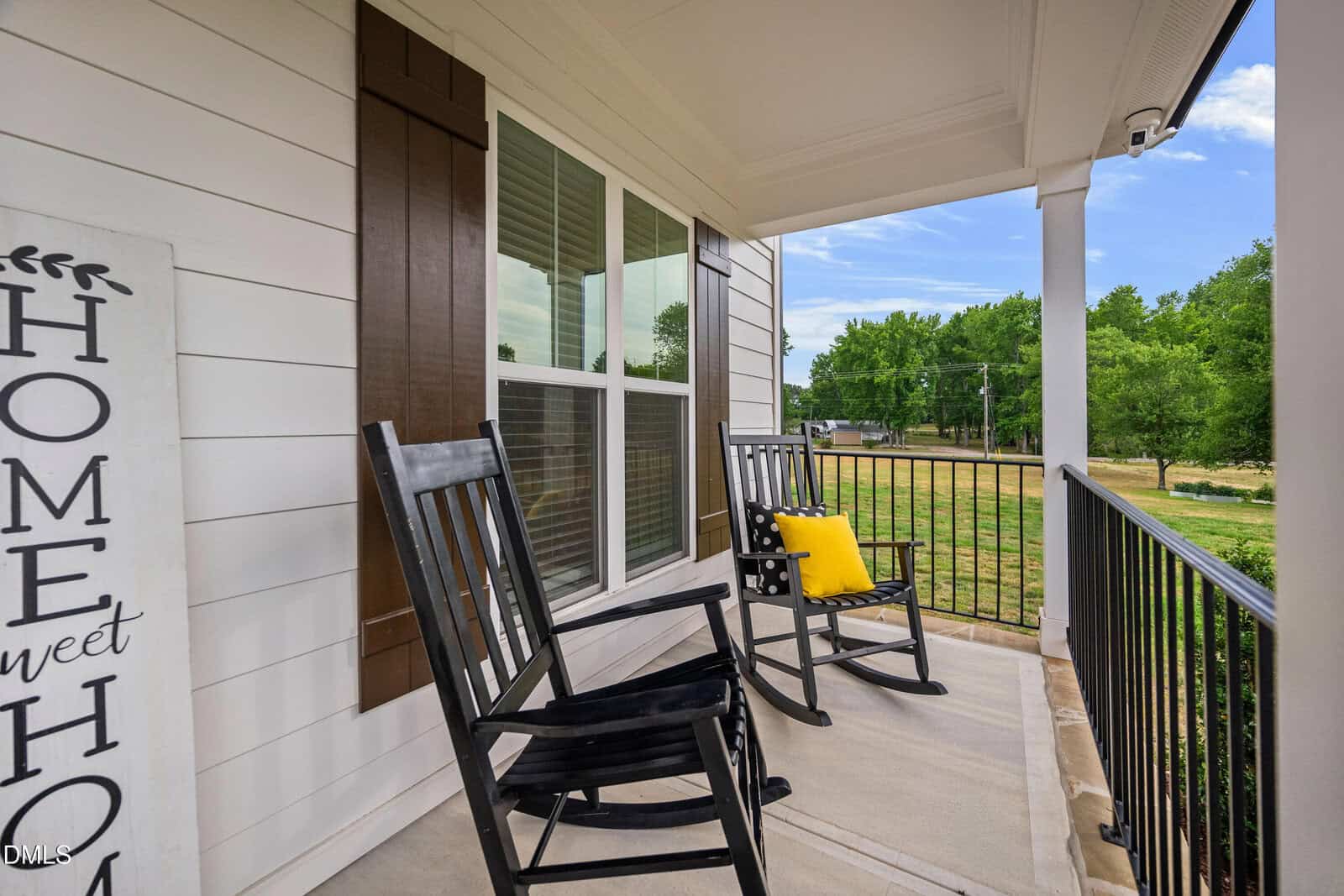 Two black rocking chairs, one with a yellow pillow, sit on a porch with white siding and brown shutters. A “Home Sweet Home” sign leans against the wall. The porch overlooks a grassy yard and trees.