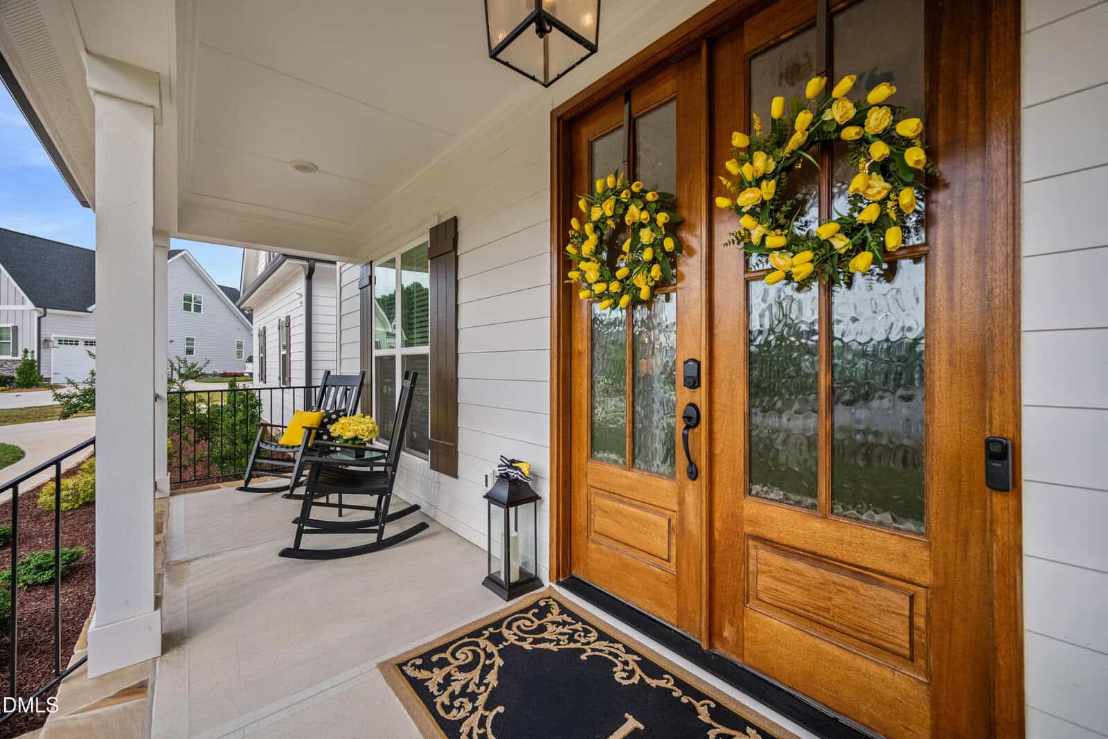 A welcoming porch with double wooden doors decorated with yellow floral wreaths, a black lantern, and two black rocking chairs with yellow cushions, set against white siding and a tidy garden.