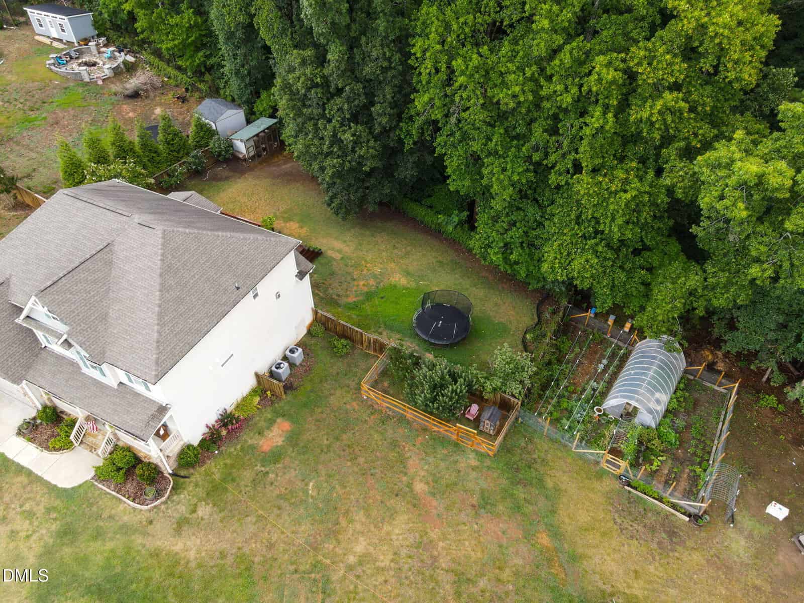Aerial view of a house with a fenced backyard, trampoline, and a garden with a greenhouse next to a wooded area. The yard has green grass and some landscaping around the house.
