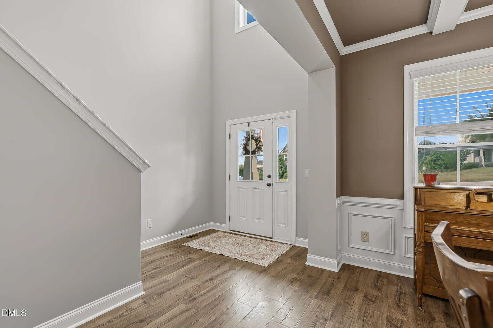 Bright entryway with wood flooring, white double doors, beige walls, a small rug, and a brown accent wall. A wooden desk sits near a window showing green grass and palm trees outside.