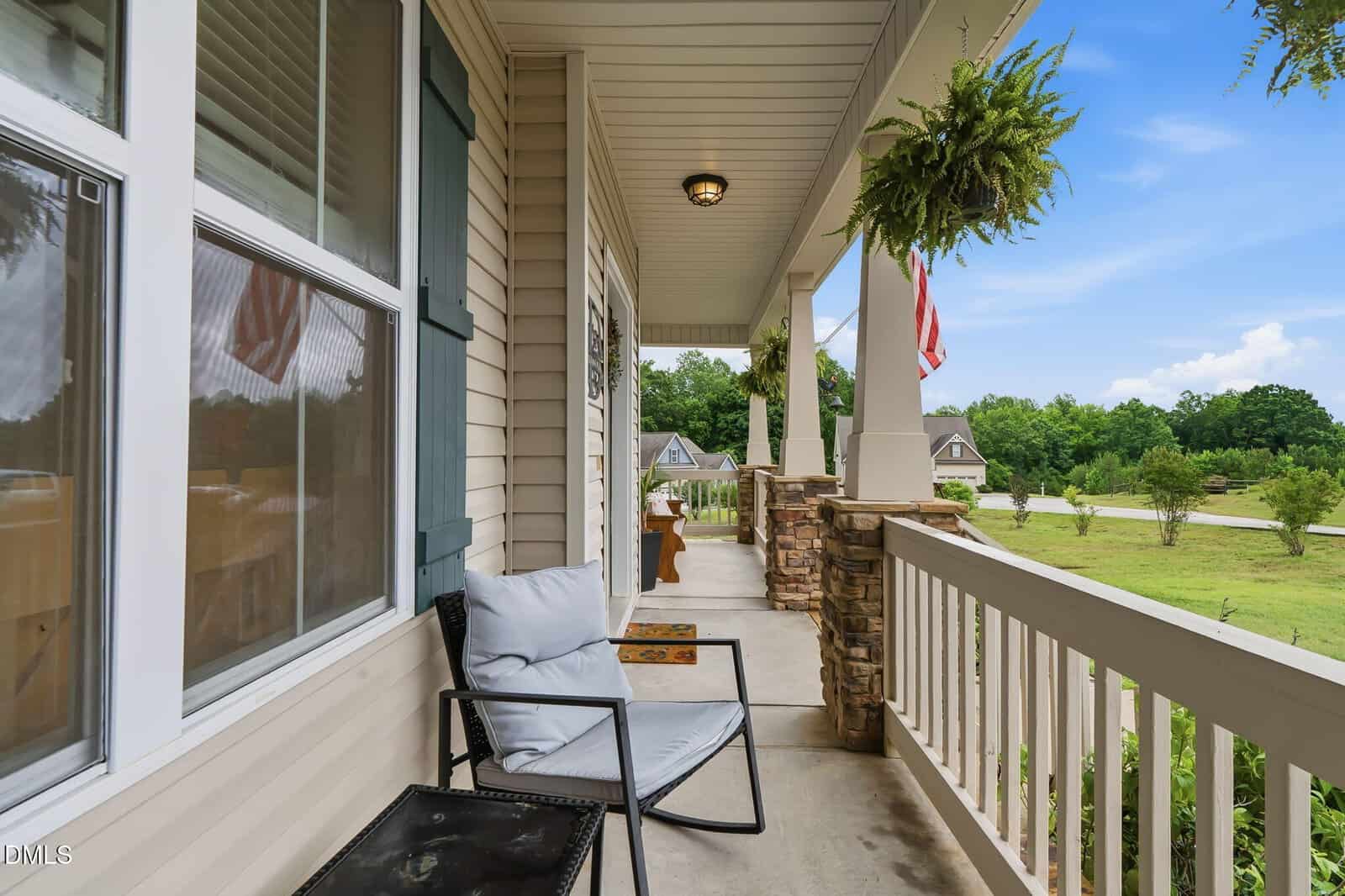 A cozy front porch with a cushioned chair, small table, hanging fern, American flag, and stone pillars overlooks a green lawn and quiet suburban street with houses and trees in the background.