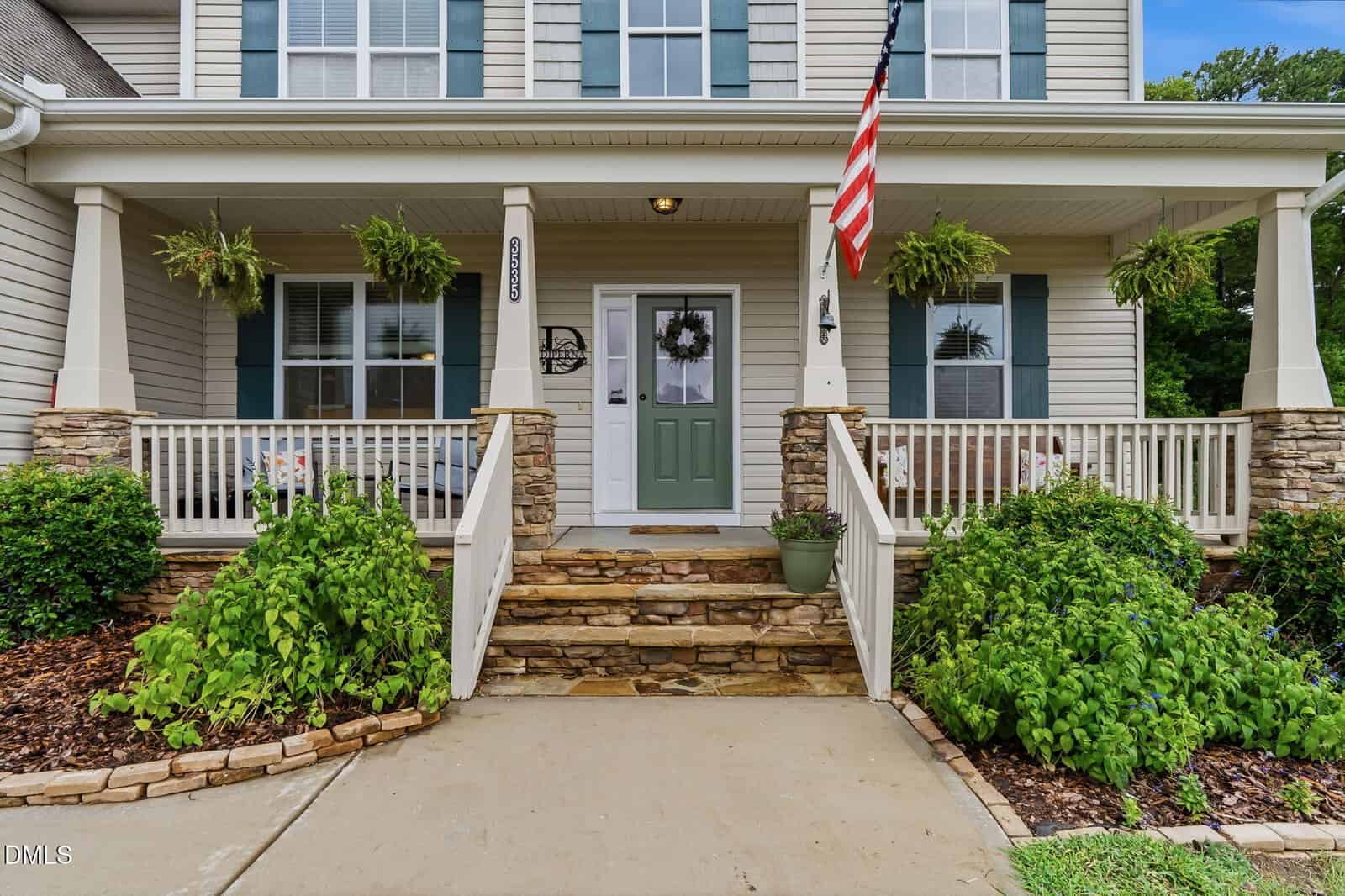 A two-story house with beige siding, stone accents, and a green front door. The porch has white railings, hanging plants, a U.S. flag, and steps leading to a landscaped walkway.