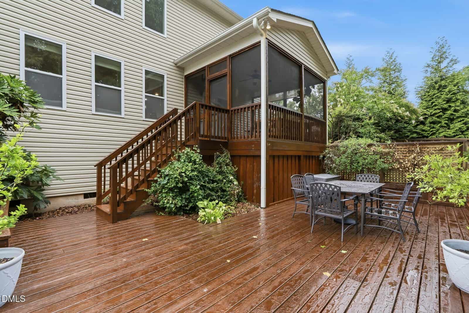 A wooden deck with patio furniture, potted plants, and stairs leading to a screened porch attached to a beige house with multiple windows. Lush greenery surrounds the outdoor space.