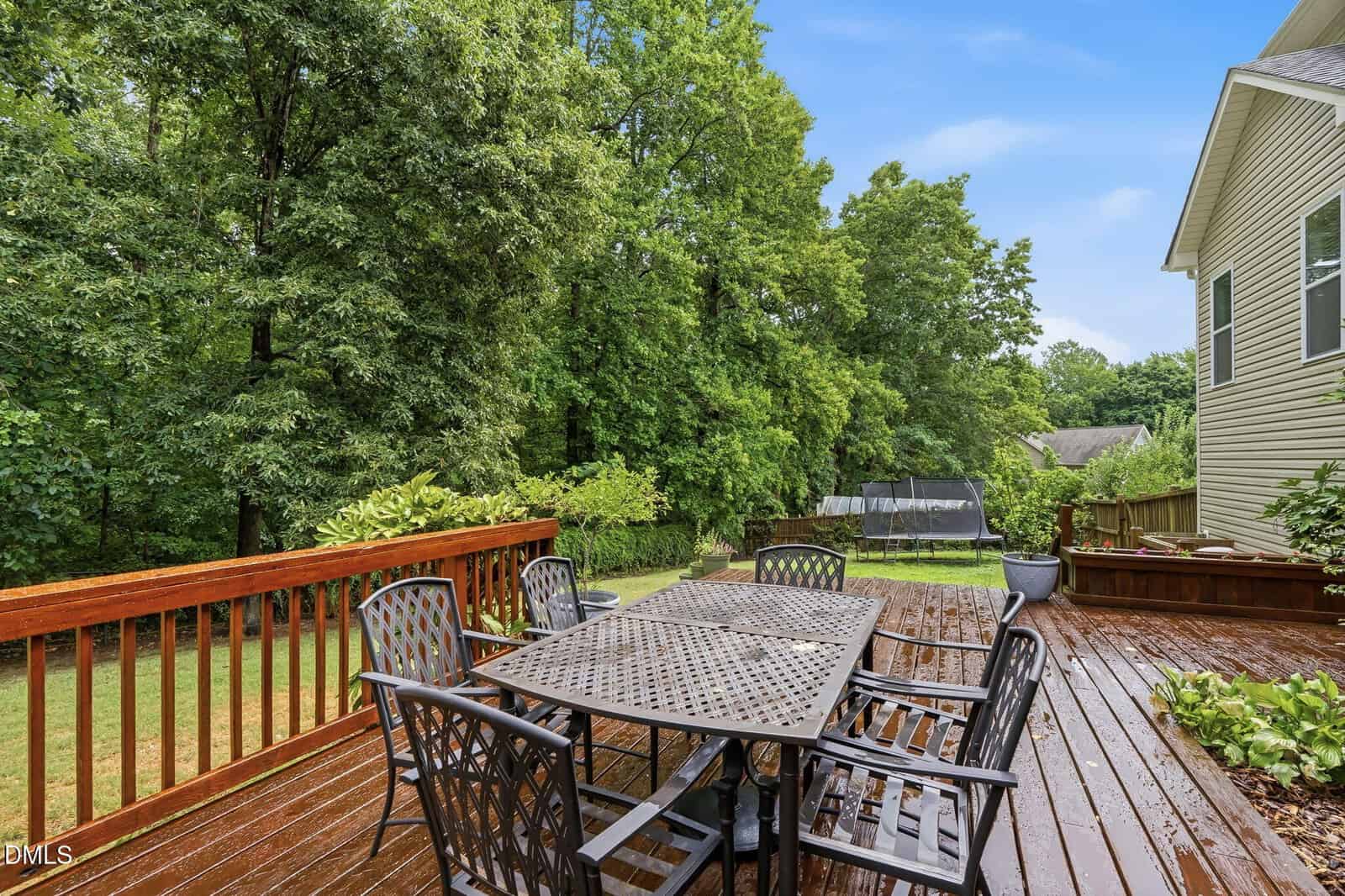 A wooden deck with a metal outdoor dining set overlooks a green backyard with trees and a trampoline, next to a house on a clear day.