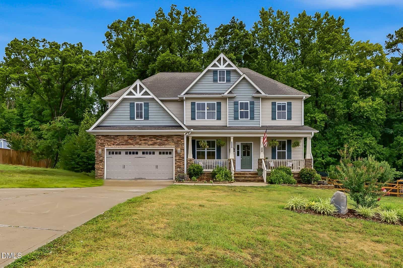 Two-story gray house with white trim, a front porch, double garage, and stone accents, set against a backdrop of tall trees and blue sky, with a green lawn and landscaped bushes in front.