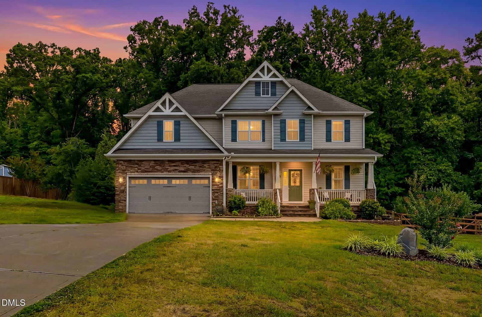 A two-story suburban house with gray siding, a front porch, and a two-car garage, set against a backdrop of tall trees and a colorful sunset sky. The lawn is green with some landscaping near the entrance.