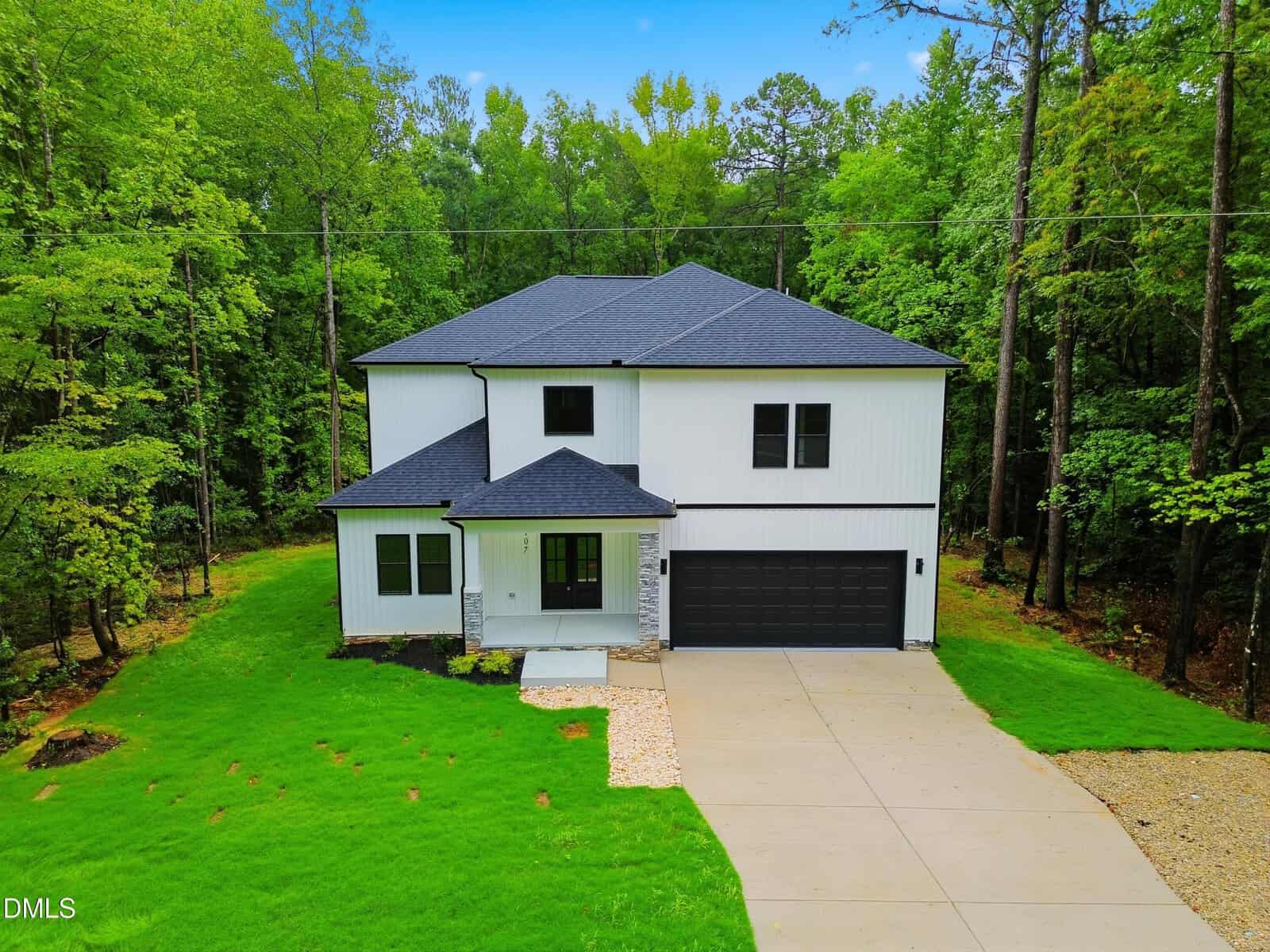 Modern two-story house with white siding and black roof, a covered front porch, attached two-car garage, and a wide driveway, surrounded by lush green lawn and trees.