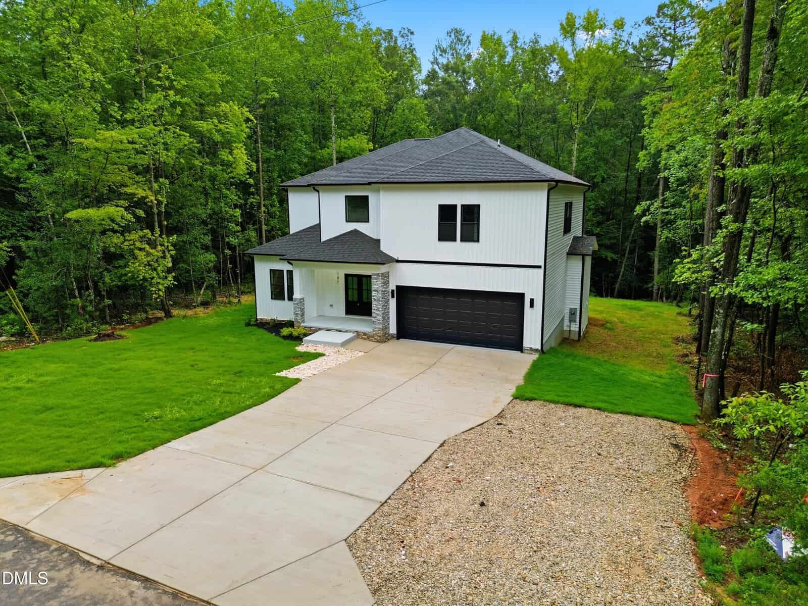 Modern two-story white house with black accents, a covered porch, and a two-car garage. Surrounded by a green lawn and trees, with a concrete driveway and gravel parking area.