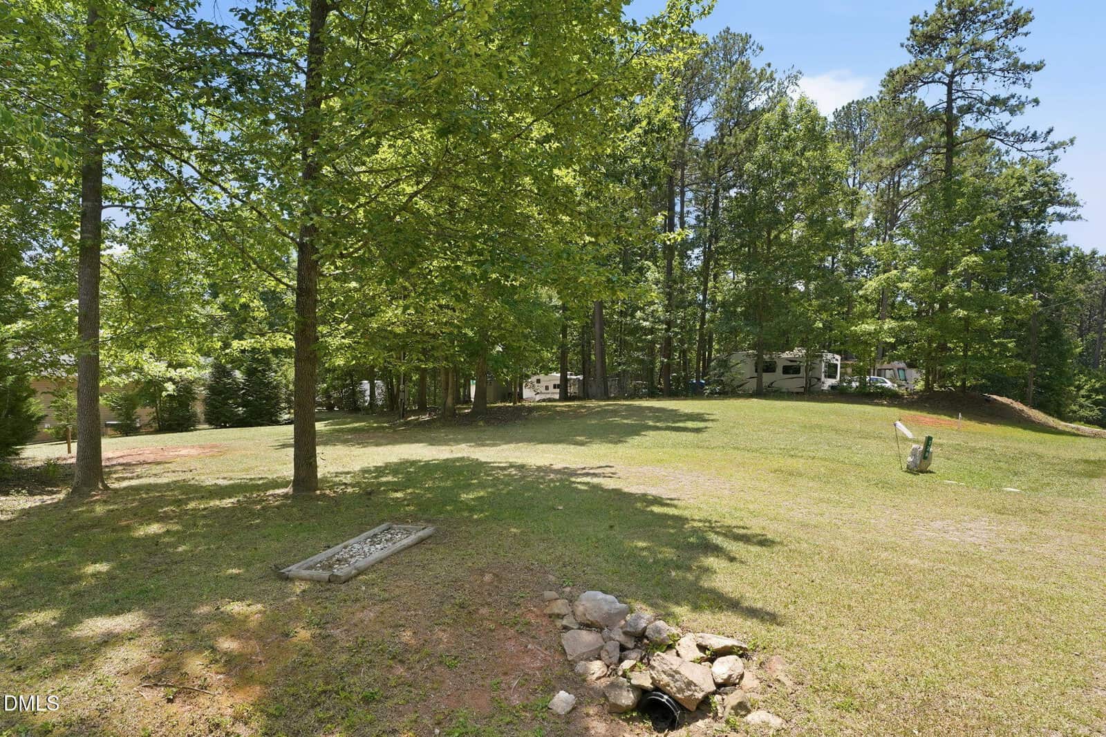 A grassy clearing with scattered trees, a small drainage area with rocks in the foreground, and an RV parked among trees in the background on a sunny day.