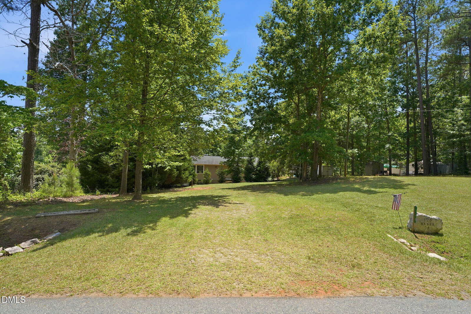 A grassy driveway leads to a house partially hidden by tall green trees. An American flag is placed beside a stone marker on the right, and the scene is surrounded by a wooded area under a clear blue sky.