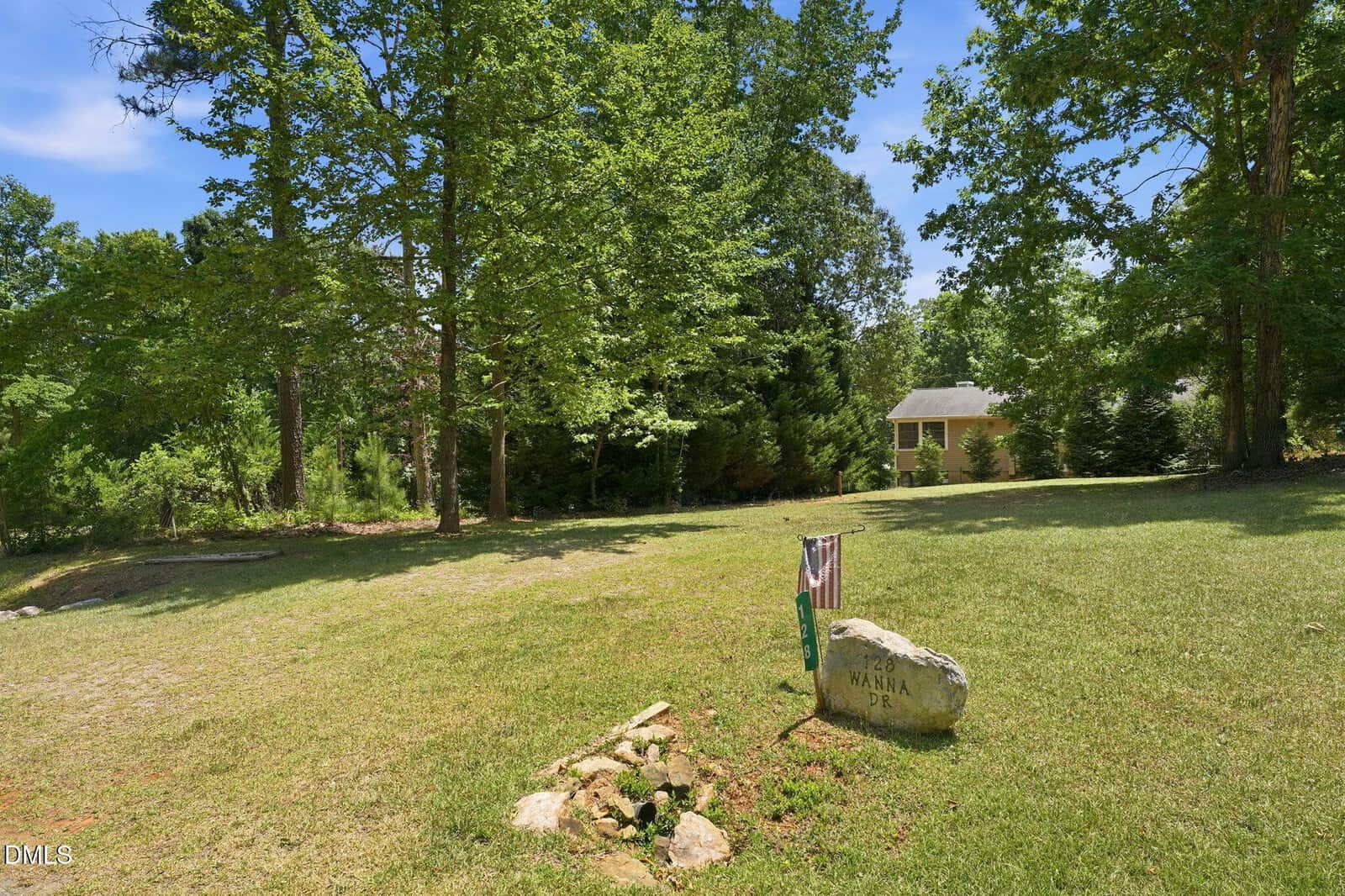 A grassy yard with scattered trees, a small American flag by a large rock labeled “WANA PR,” and a house partially visible in the background, surrounded by greenery.
