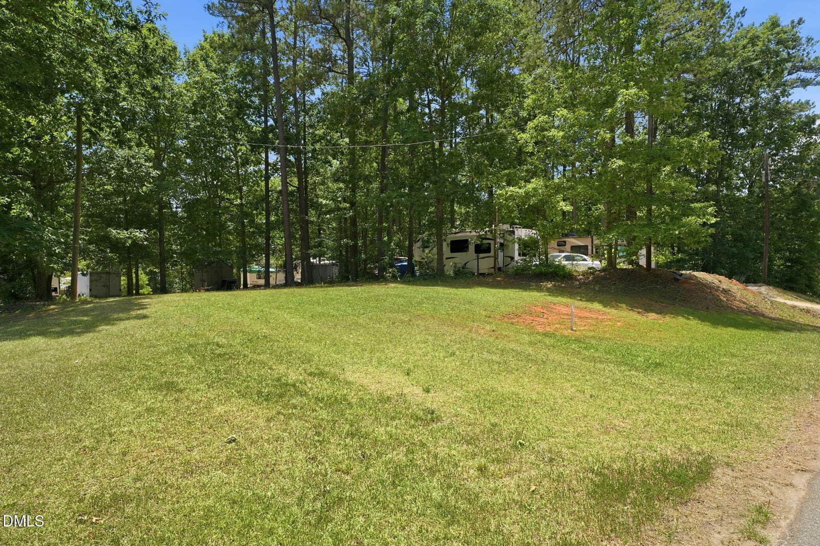 A grassy hill slopes up to a wooded area with several RVs parked among tall trees under a clear blue sky.
