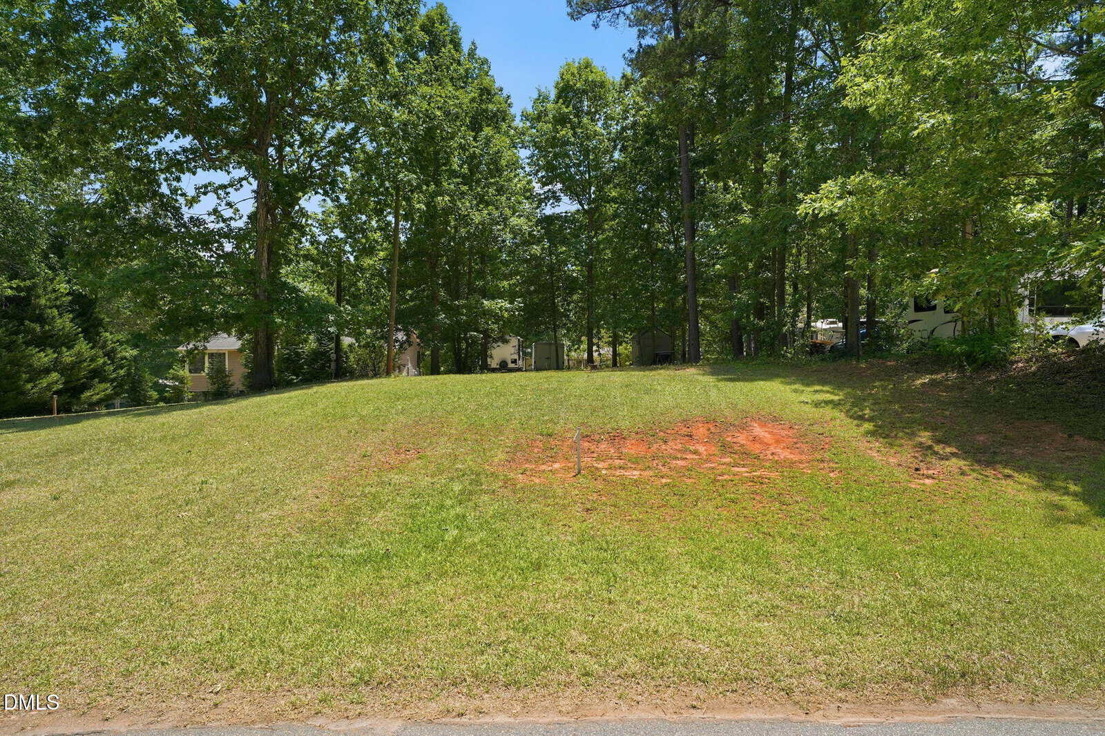 A grassy, slightly sloped vacant lot with patches of bare reddish soil, bordered by tall green trees and partially shaded, on a sunny day. Houses are visible in the background through the trees.