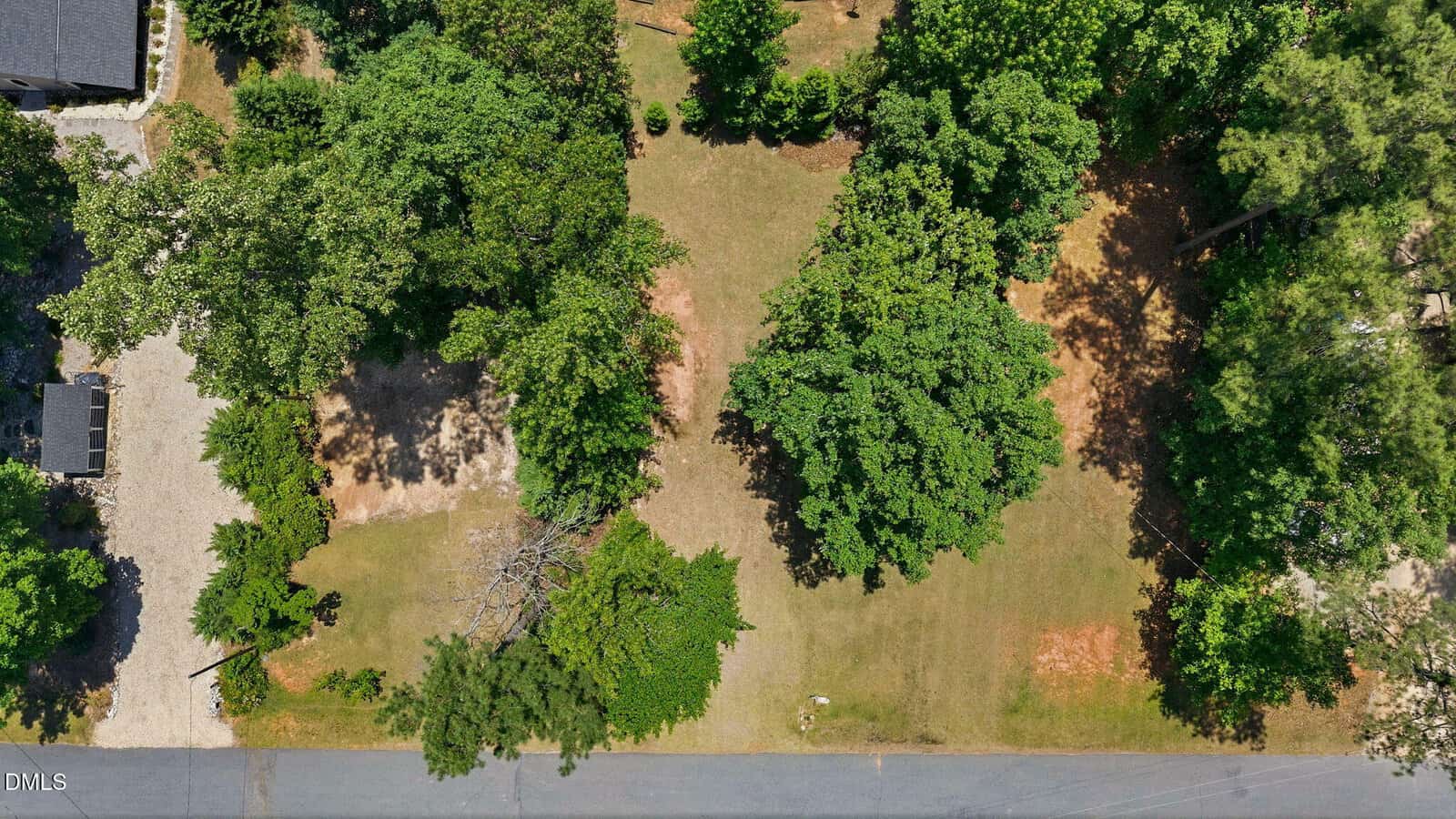 Aerial view of a grassy lot with scattered trees, a paved road at the bottom, and a gravel driveway on the left. Shadows from the trees are visible, and some bare patches of ground appear among the greenery.