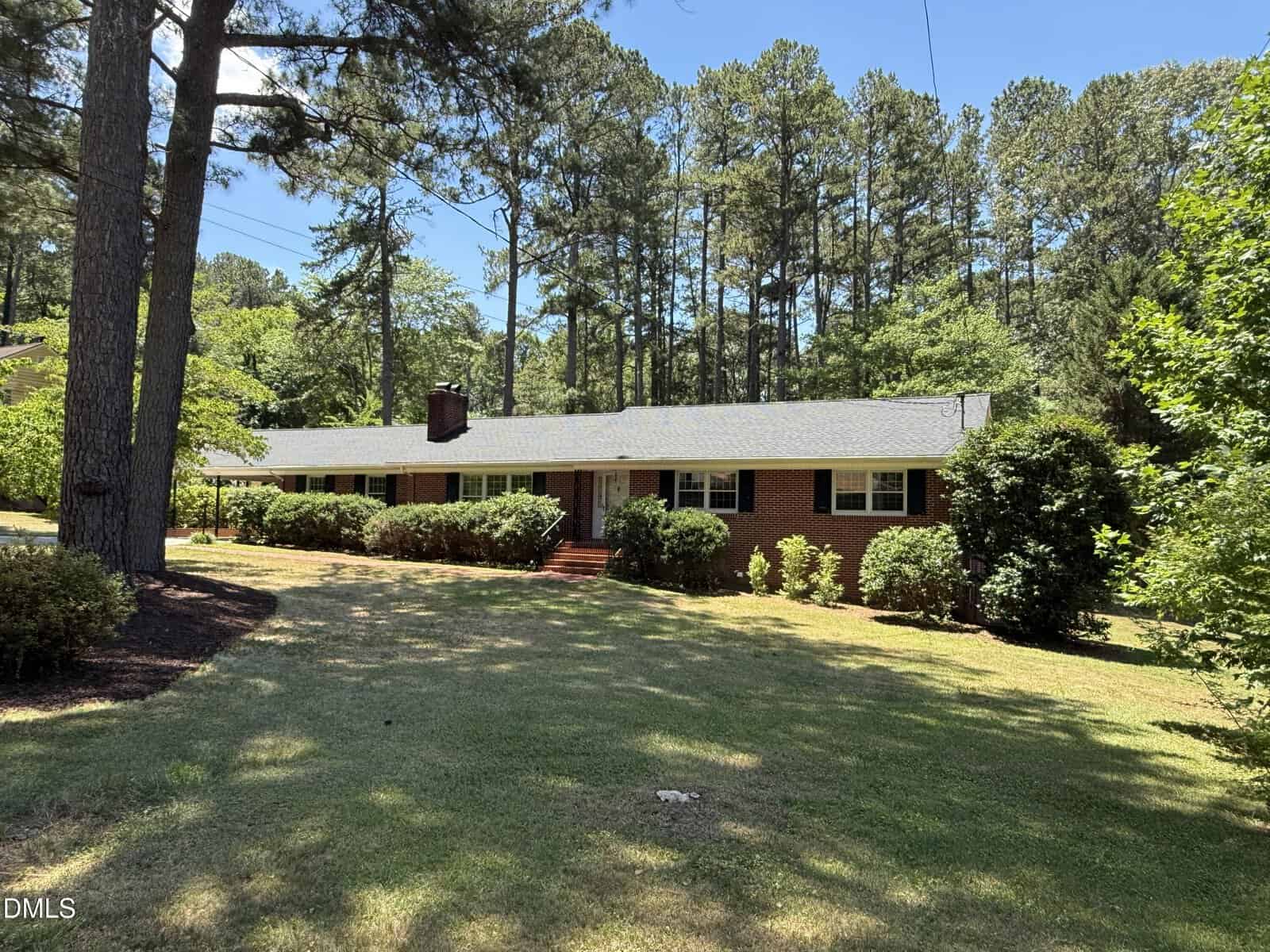 A one-story brick house with a dark roof, white trim, and multiple windows is surrounded by trees and bushes on a grassy lawn under a clear blue sky.