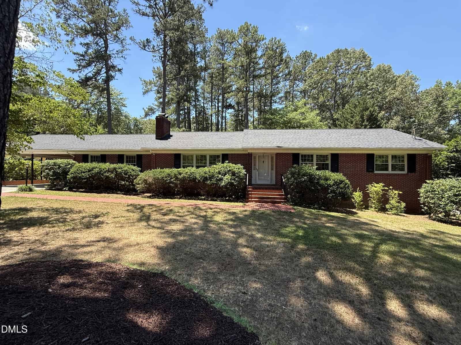 A single-story brick house with white trim at 174 Pine Cone Drive, Oxford, nestles among bushes and tall pine trees under a clear blue sky. The front yard is partly shaded, with grassy patches and a brick walkway leading to the door.