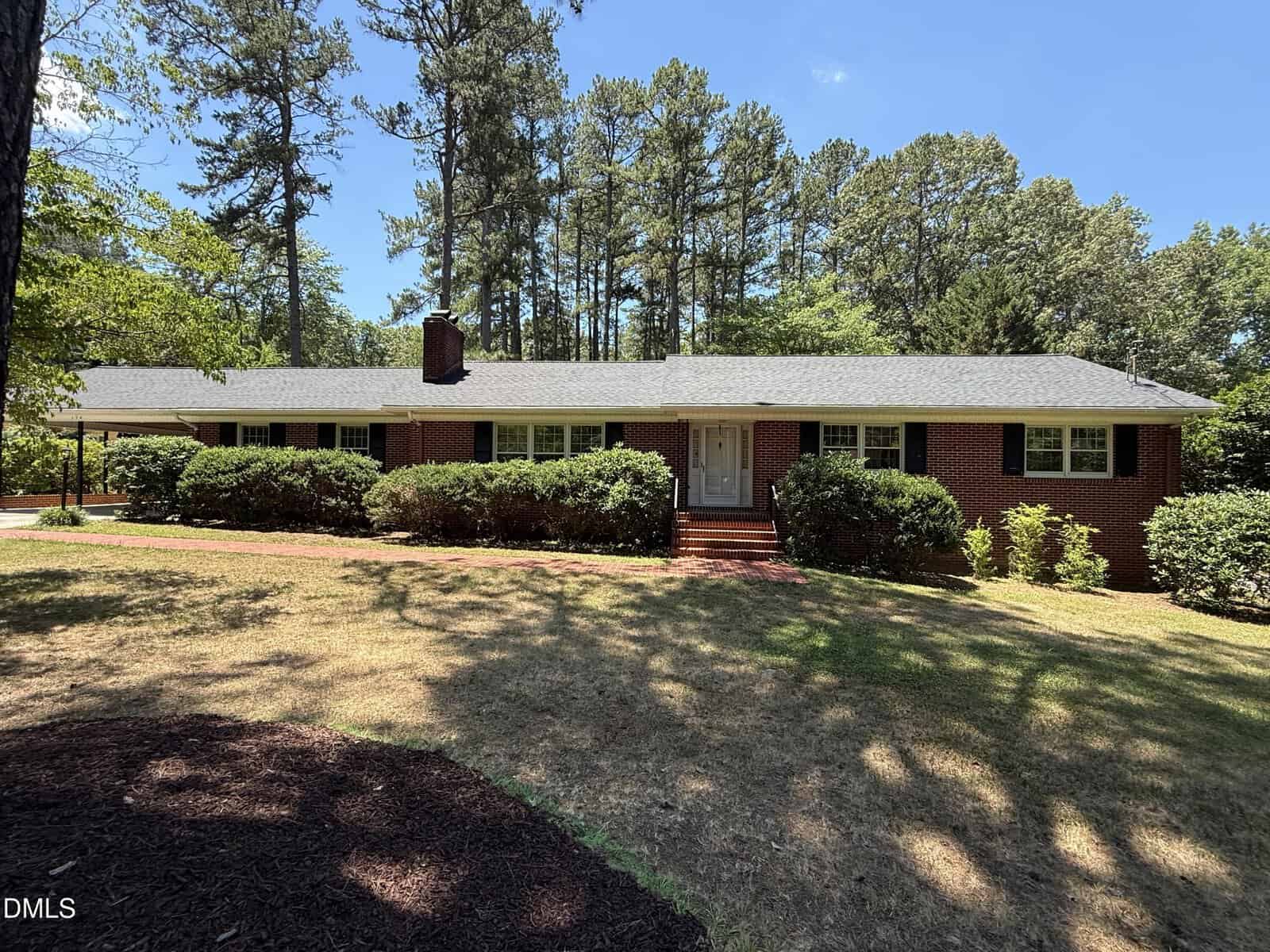 A single-story red brick house with white trim, a gray shingle roof, and green shutters, surrounded by shrubs and tall pine trees under a clear blue sky.