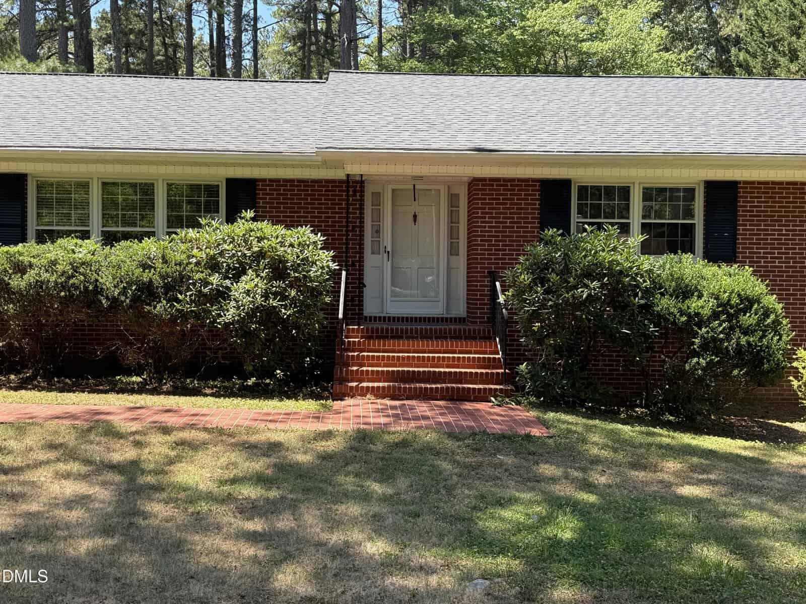 A brick house with white trim and a gray shingled roof, featuring a white front door, black shutters, red brick steps, and green bushes lining the front. Tall trees are visible in the background.