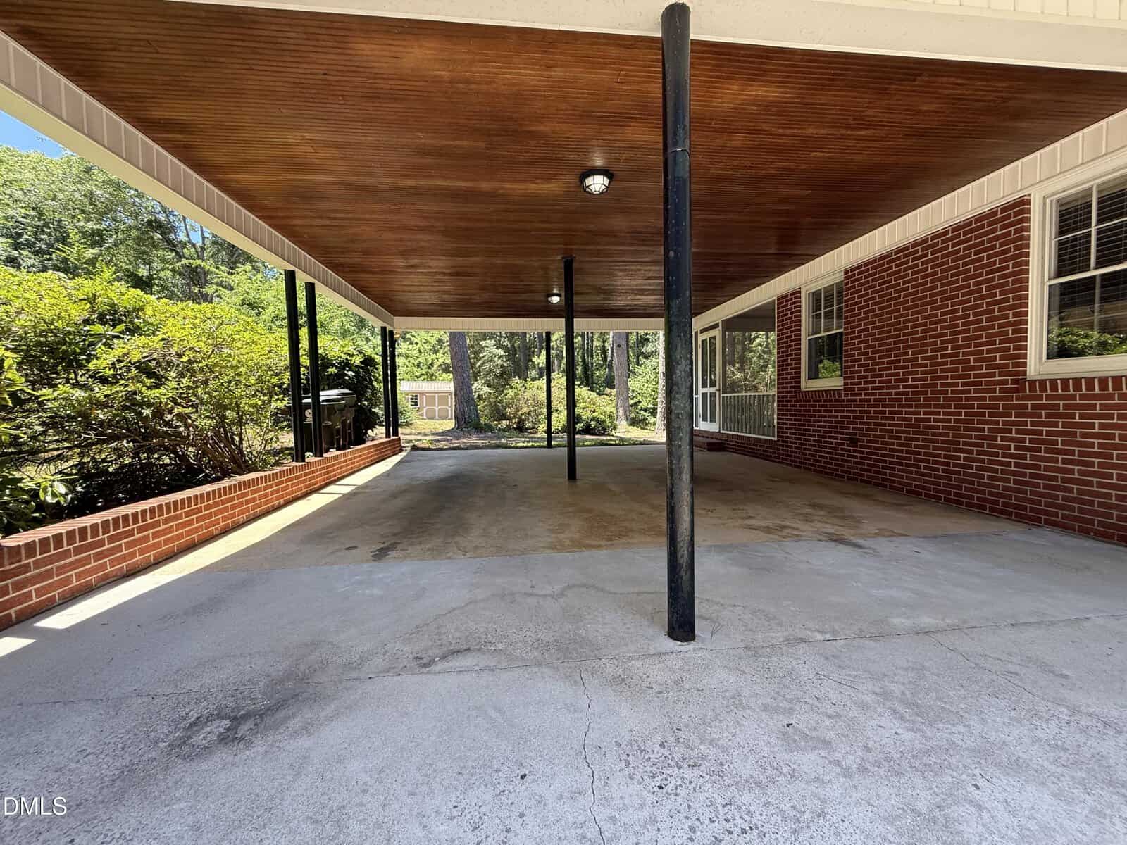 A covered carport with a wooden ceiling, black support poles, and concrete floor attached to a brick house. Green bushes line the left side, and a yard with trees is visible in the background.