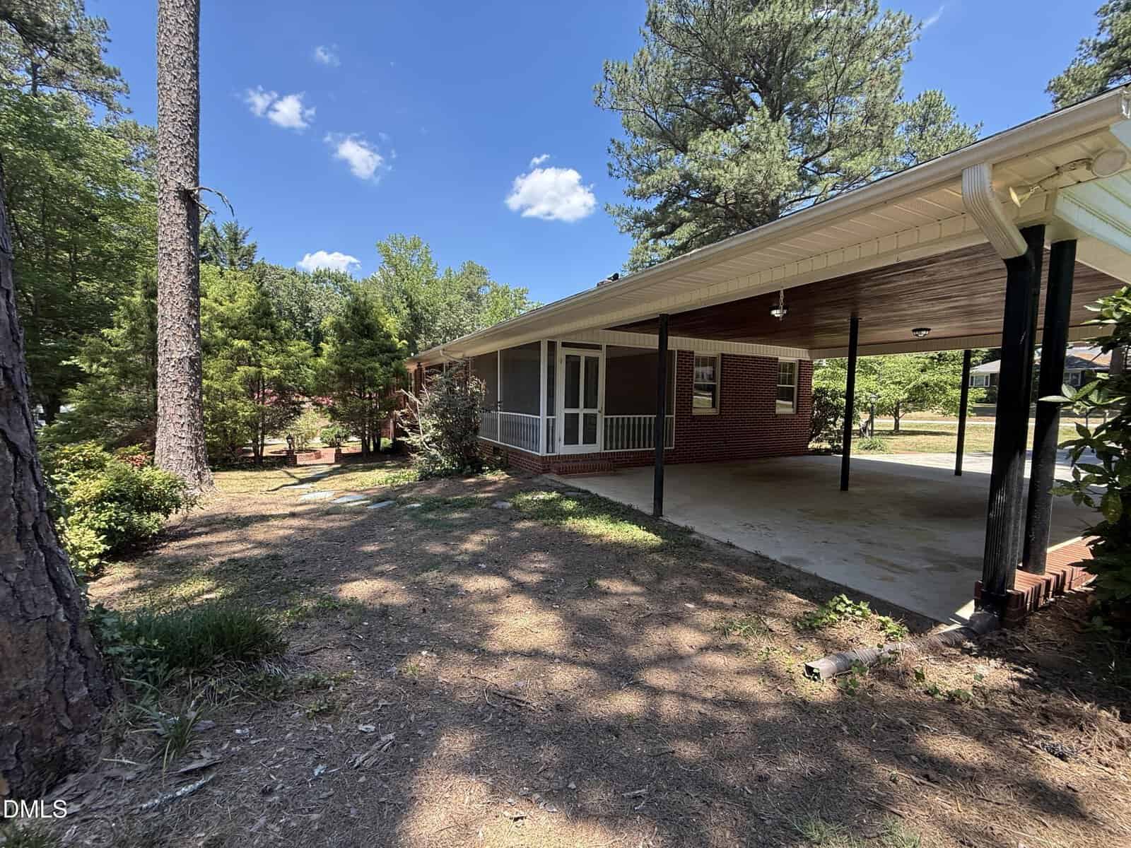 A red brick house with a covered carport, white trim, and a screened porch, surrounded by tall pine trees and greenery under a sunny blue sky. Dirt and patchy grass cover the foreground.