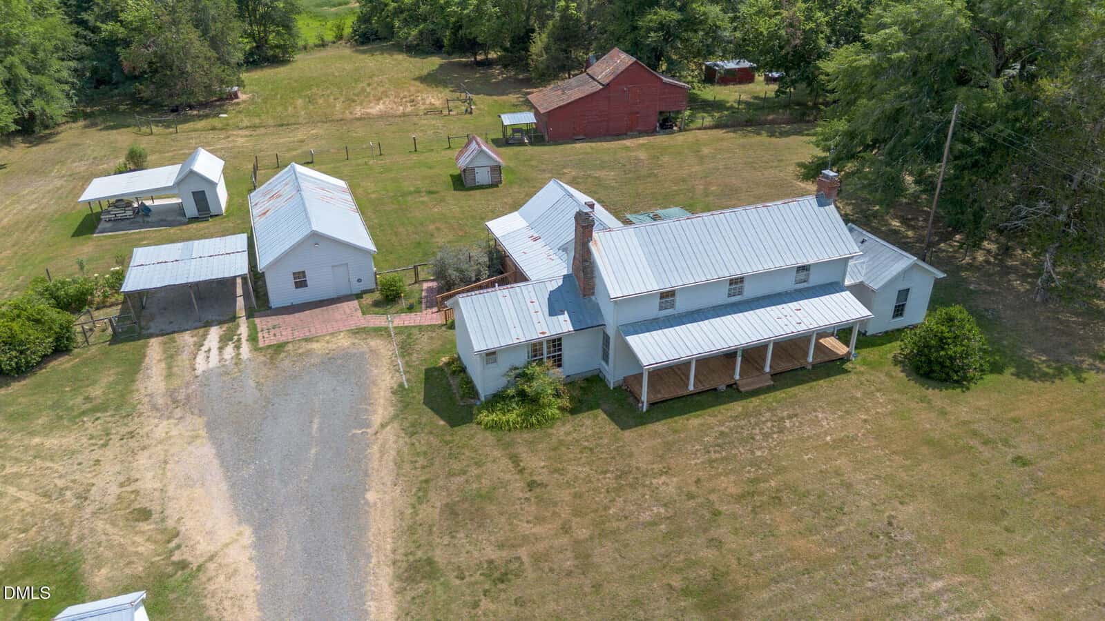 Aerial view of a rural property with a white farmhouse, multiple outbuildings, a gravel driveway, and green trees and grass in the background. The roofs are metal and the main house has a covered porch.