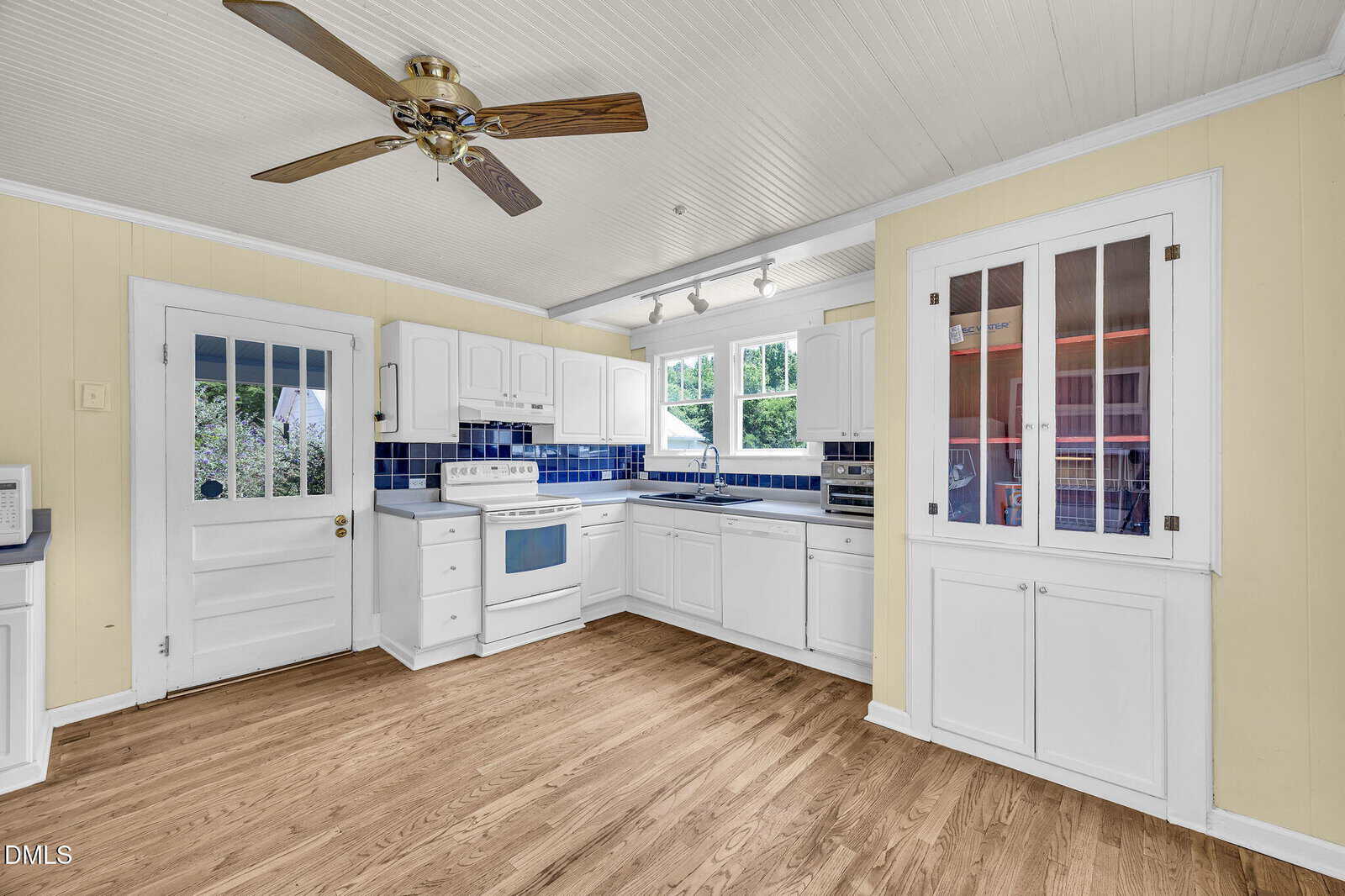 Bright kitchen with white cabinets, blue tile backsplash, white appliances, wood flooring, a ceiling fan, large windows over the sink, and a glass-front cabinet. A door leads outside, and track lighting illuminates the workspace.