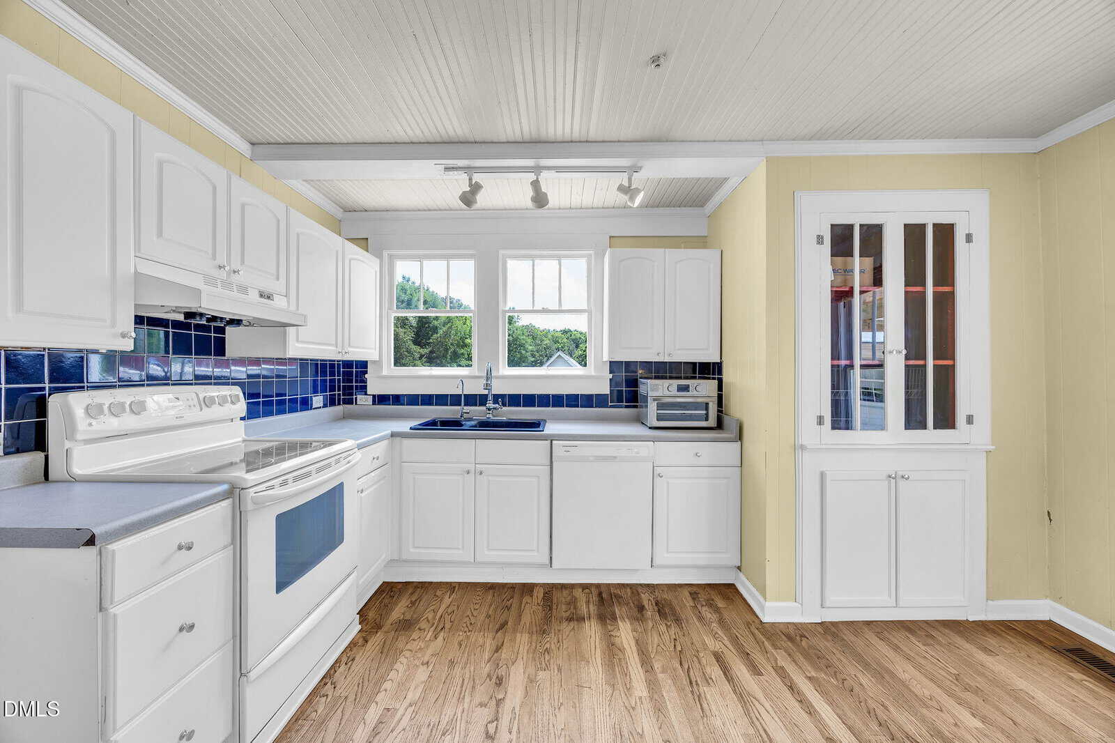 Bright kitchen with white cabinets, blue tile backsplash, white appliances, wood floor, large windows above the sink, and built-in glass-front cabinet on the right. Yellow walls and white ceiling with track lighting.