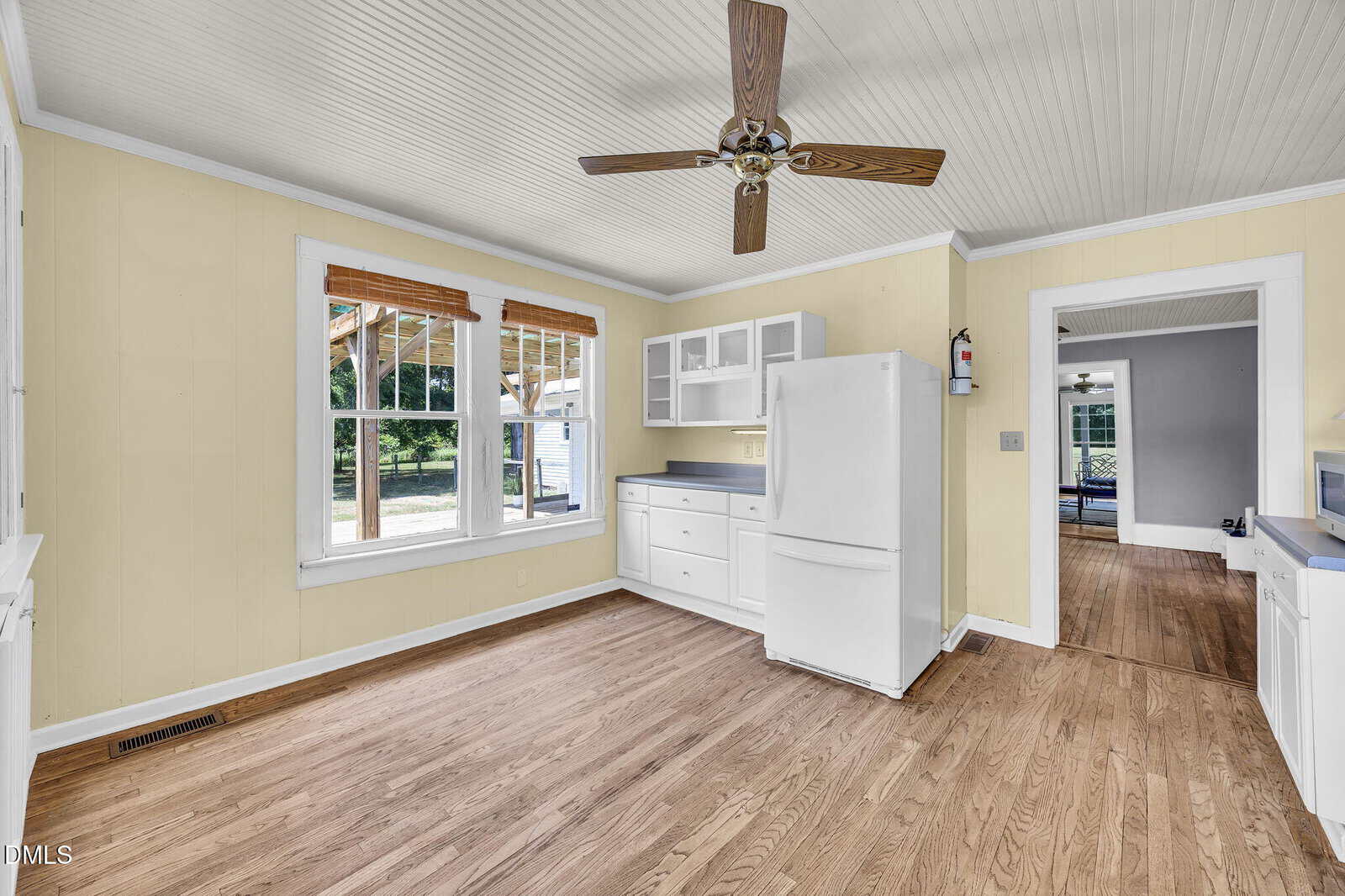 Bright kitchen with light yellow walls, wood flooring, white cabinets, white refrigerator, ceiling fan, and large windows letting in natural light. An adjacent room with hardwood floors is visible through the doorway.
