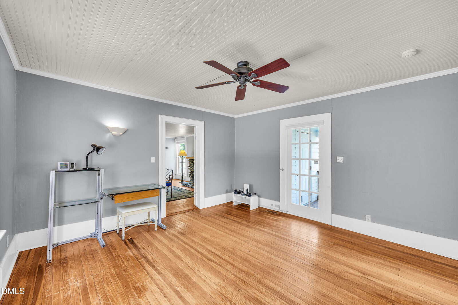 A spacious room with gray walls, wood flooring, white trim, a ceiling fan, a small desk with a lamp, and a door with glass panels leading to another area.