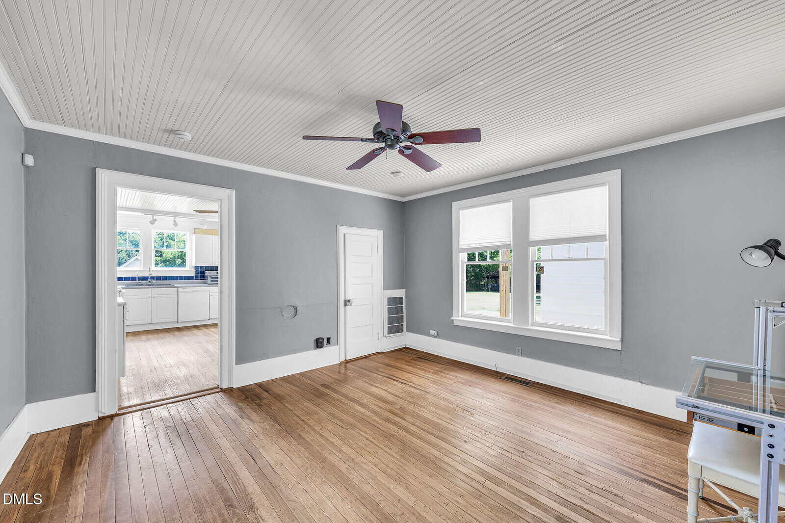 A bright room with gray walls, wood floors, a red ceiling fan, large windows, and a doorway leading to a white kitchen with more windows.