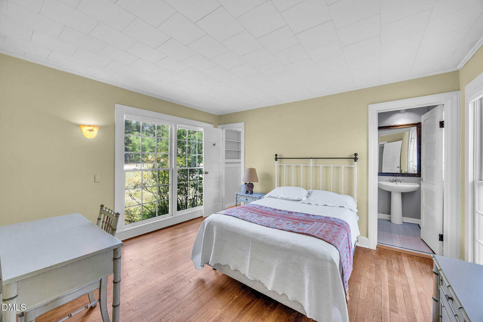Bright bedroom with wood floors, a double bed with a white metal frame, a desk with a chair, large windows overlooking greenery, and an ensuite bathroom with a pedestal sink and mirror.