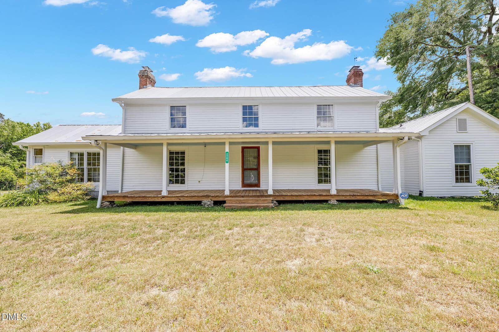 A white two-story farmhouse with a metal roof, wooden steps, and a large covered front porch sits on a grassy lawn under a blue sky, just outside Oxford at 2204 US 158.