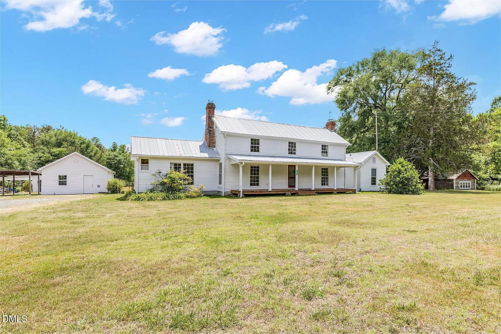 A white two-story farmhouse with a metal roof and brick chimneys sits on a large, grassy lawn at 2204 US 158, Oxford. Trees and a detached building are visible in the background under a blue sky with scattered clouds.