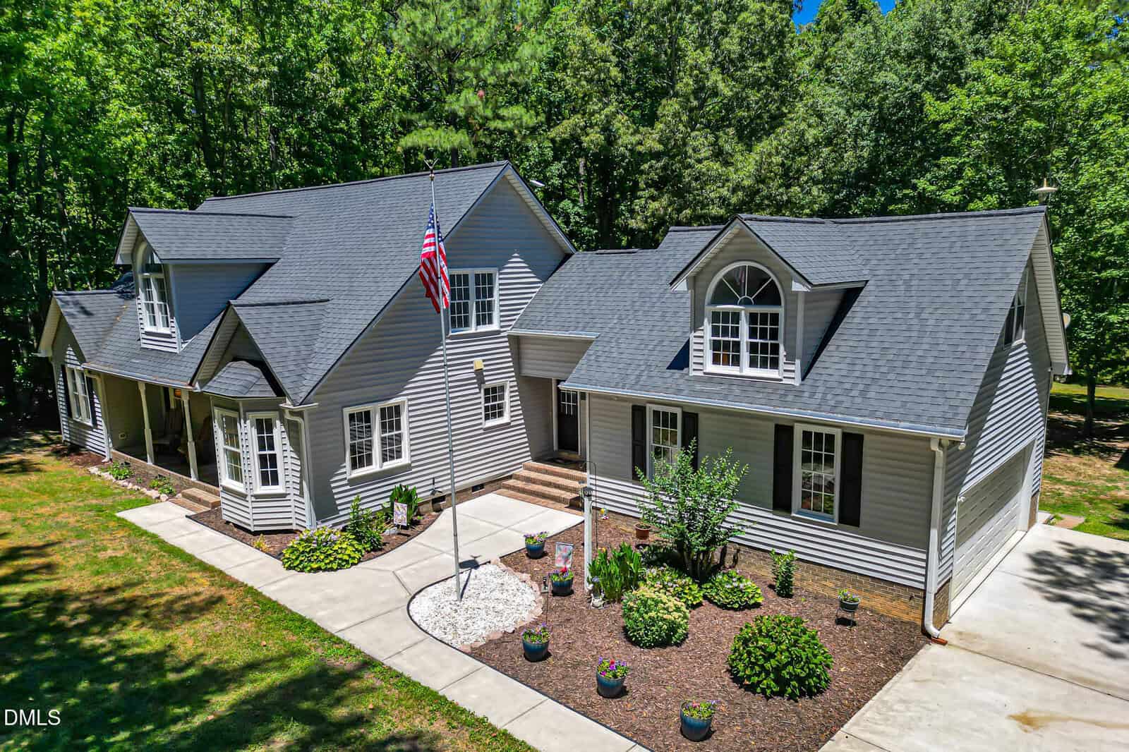 A two-story suburban house with light gray siding, dark gray roof, dormer windows, an American flag, a front porch, landscaped garden beds, and a concrete driveway, surrounded by trees.