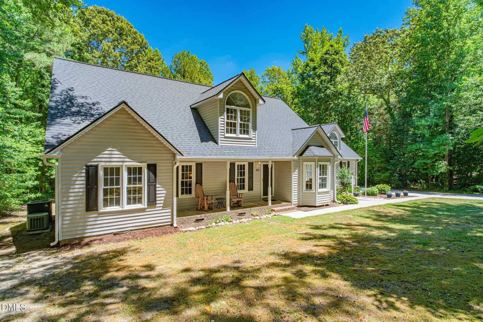 A beige house with a dark gray roof and dormer window sits among trees. A porch with chairs is at the front, and an American flag is displayed on a pole by the driveway on a sunny day.